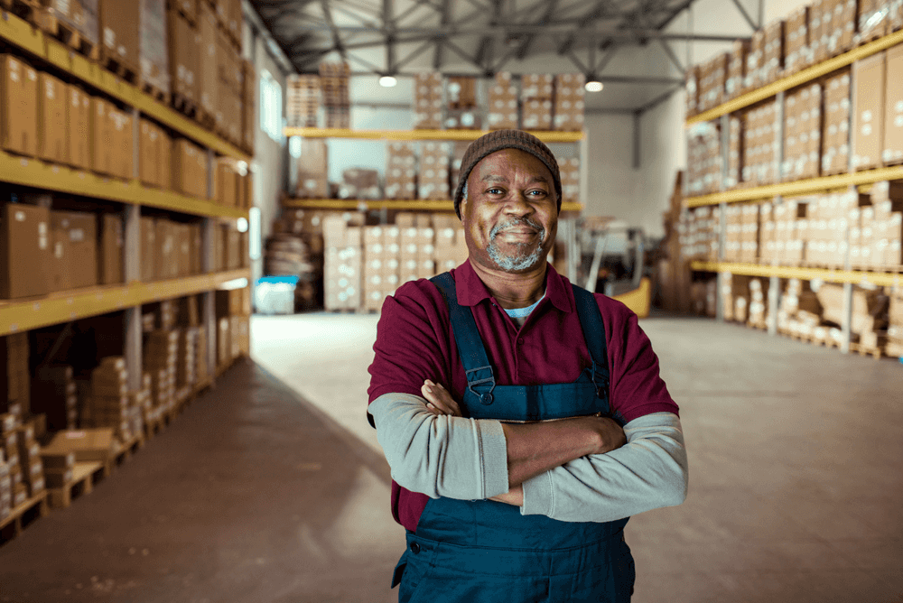 A man smiling towards the camera with a humble but pleased kind of look, his arms crossed, standing in a warehouse