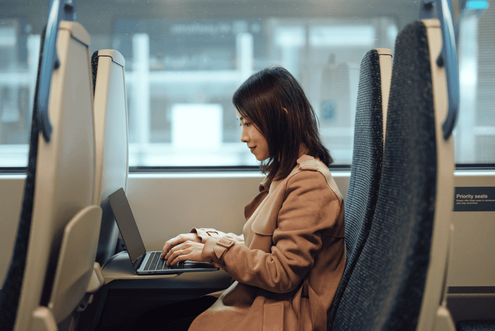 A woman is sitting comfortably on a brightly lit commuter train and working on her laptop