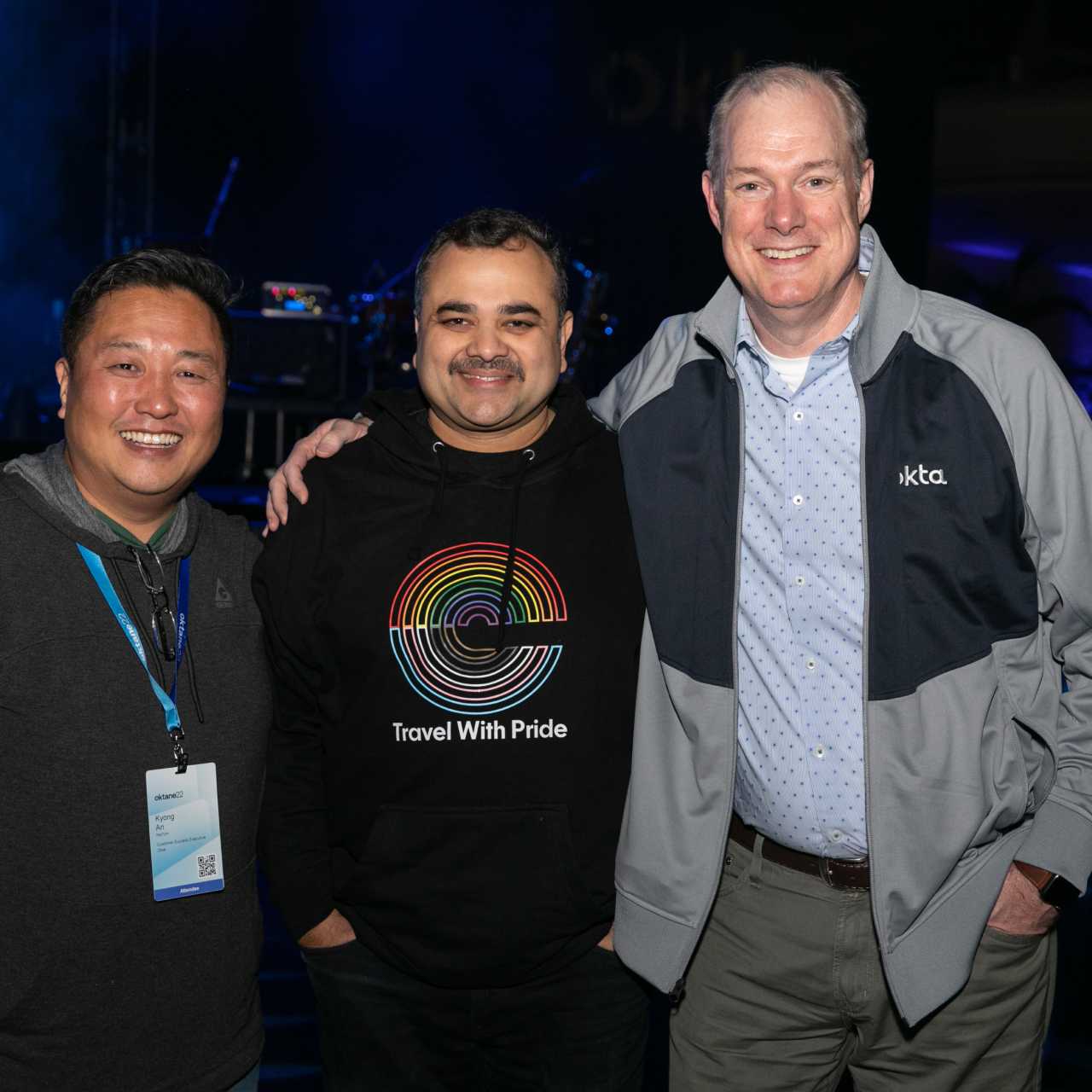 Three men posing for a photo while one man wears a Travel with Pride Okta t-shirt.