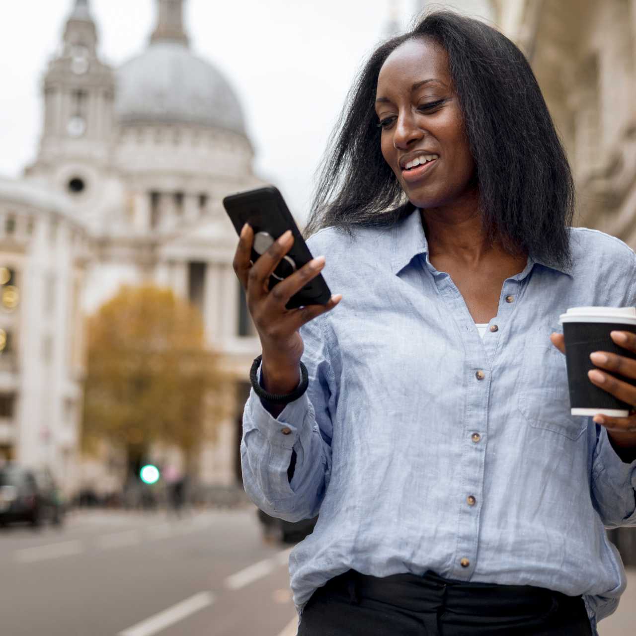 Woman wearing a buttoned blouse walking down a city street with a government building in the background with coffee in one hand and smart phone in the other. 