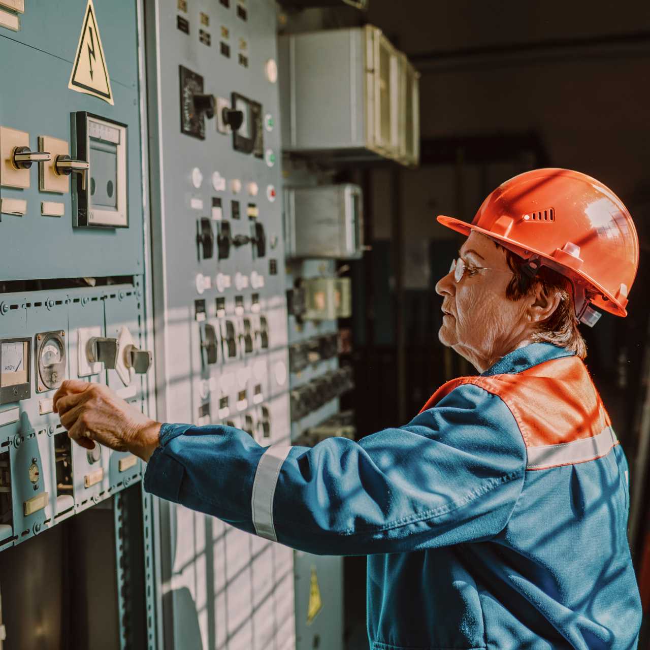 Older woman wearing blue and orange uniform and orange hard hat in control room touching control buttons.