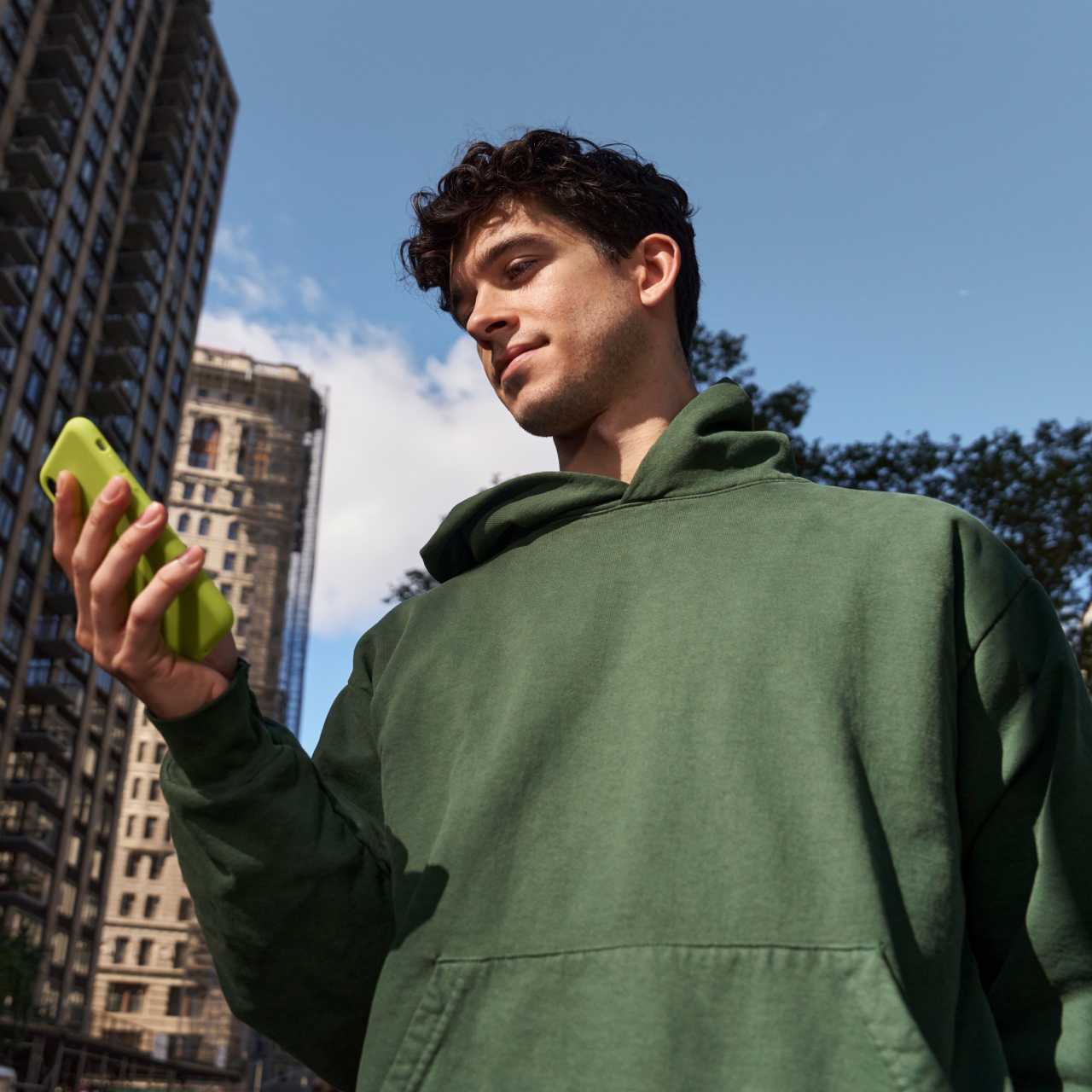 Young man in a green sweatshirt standing on city street while looking at lime green smartphone