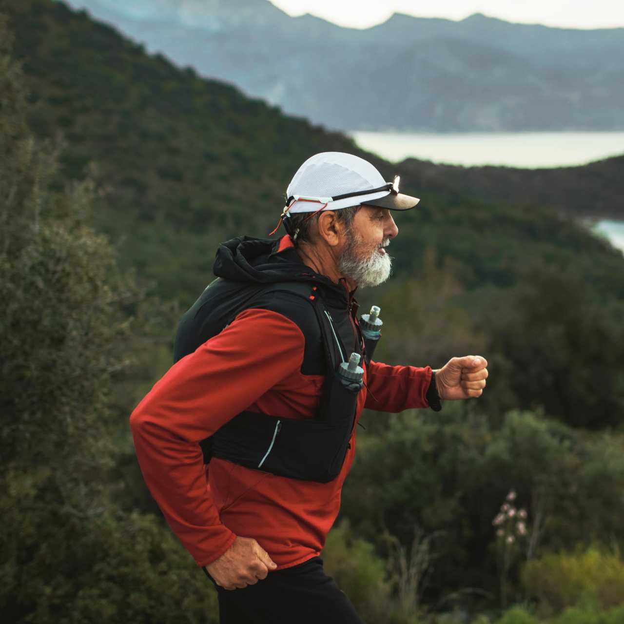 Older man in red shirt, black running vest, and white hat running through mountainous trail