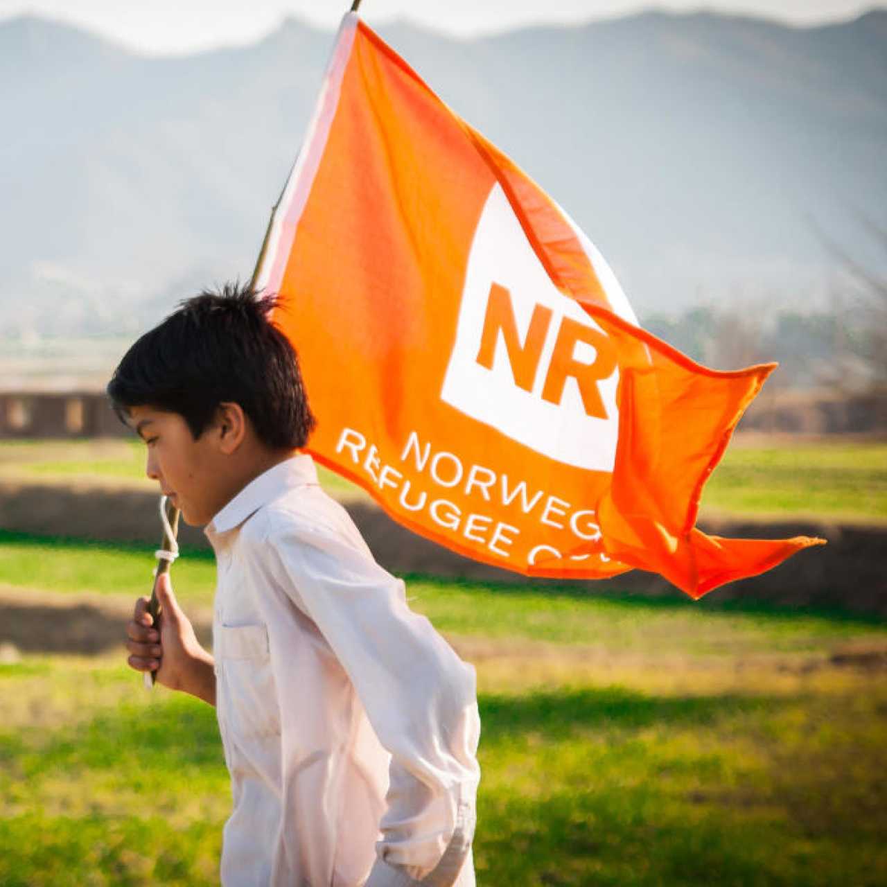 Child running in green field while holding orange Norwegian Refugee Council flag