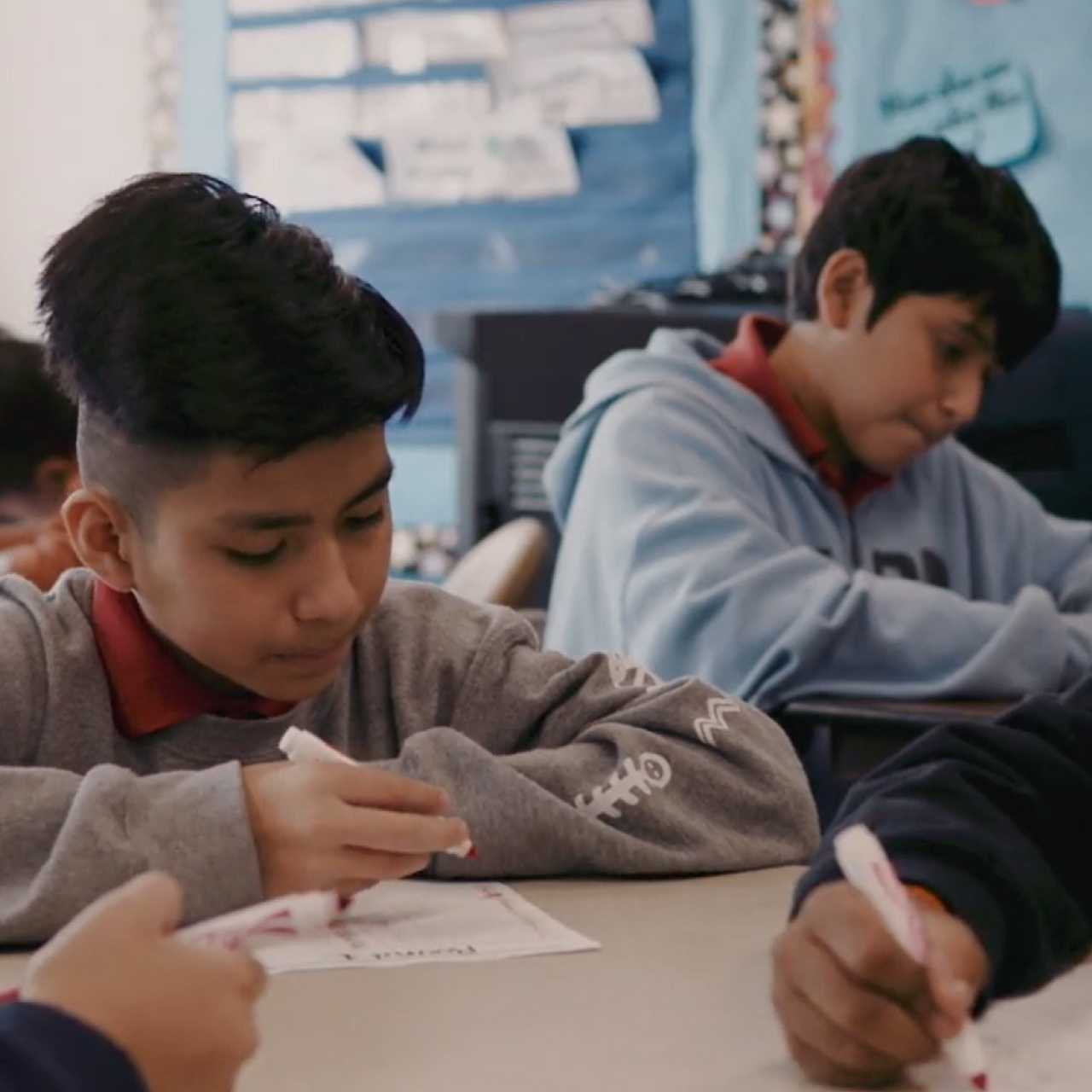Two boys in an elementary school classroom sitting at desks while writing on paper with markers