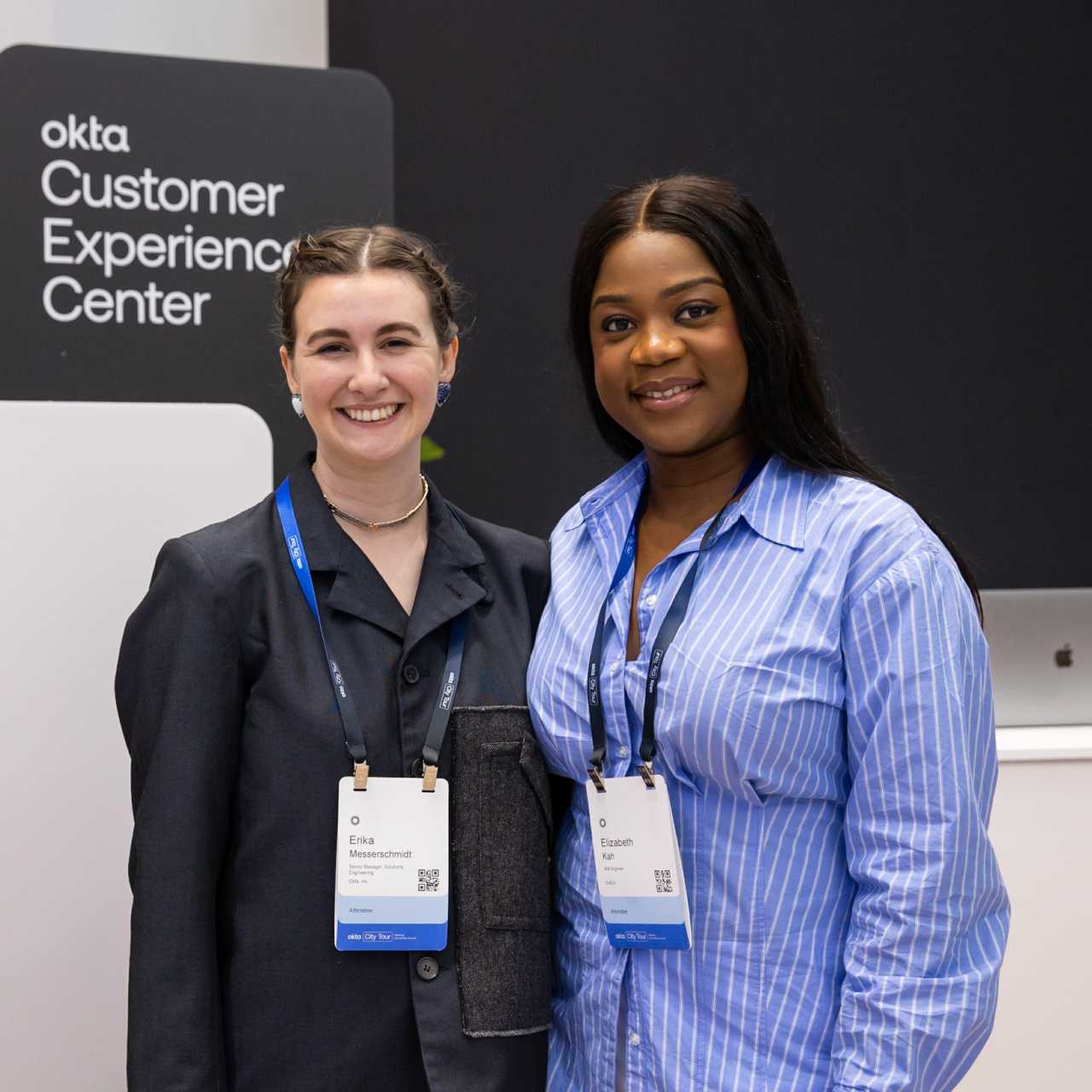 Two female employees posing next to Okta Customer Experience center sign