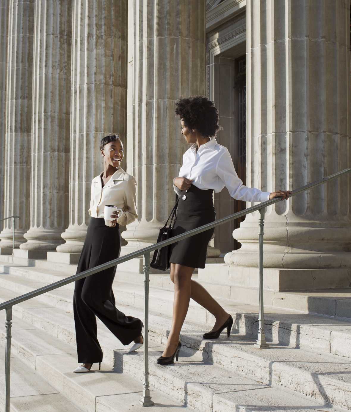 Two women in professional clothes, taking and smiling while walking down government building steps.
