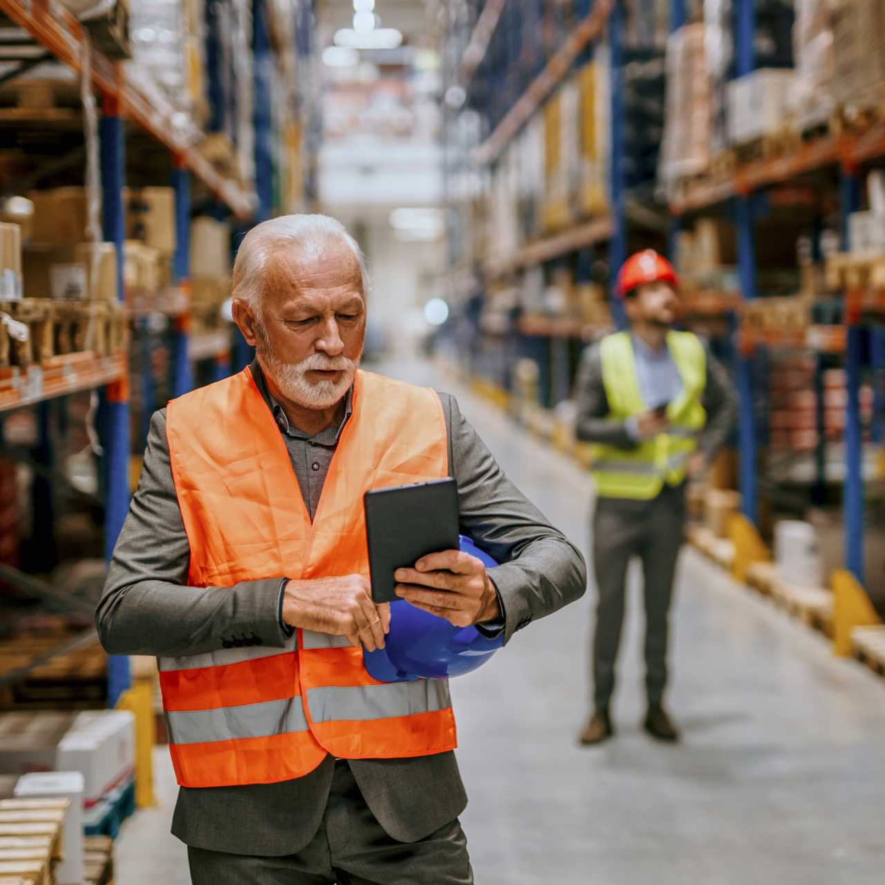 Older man wearing orange vest walking through storage warehouse while looking at smart device