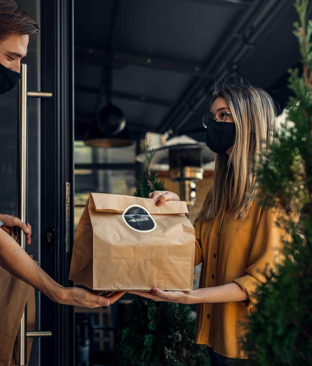 Delivery woman in mask handing a brown paper bag to a man also wearing a mask.