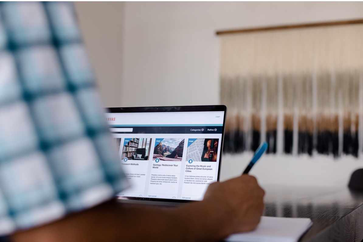 Close-up over-the-shoulder view of a person using a laptop to browse educational courses on the Canvas LMS platform while writing in a notebook.
