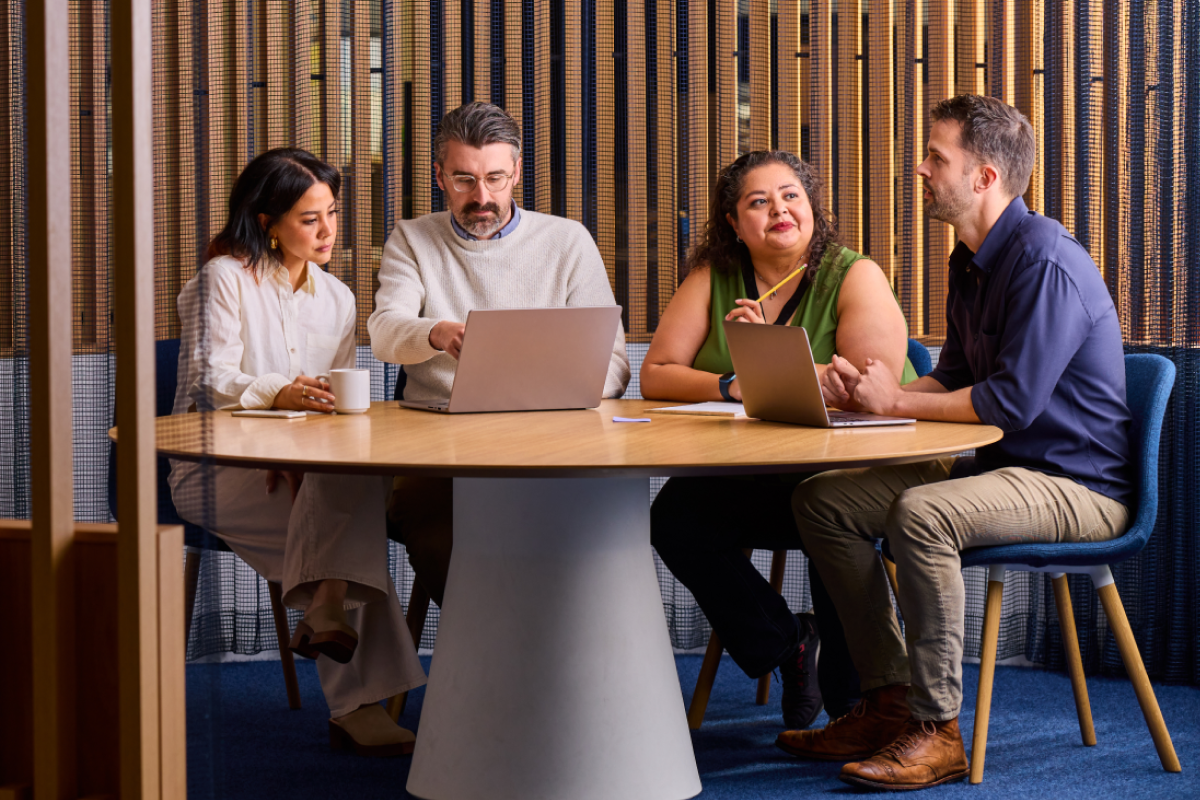 A diverse group of four colleagues sitting around a round wooden table in a modern office with slatted wood partitions, looking at a laptop and discussing a project.