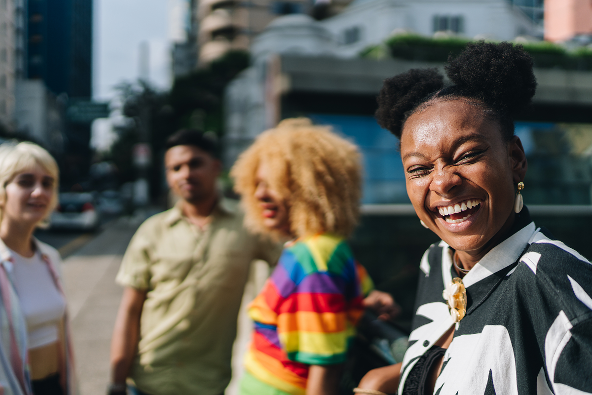 A close-up of a joyful Black woman with space buns laughing heartily, with a diverse group of three friends blurred in the background on a sunny city street.