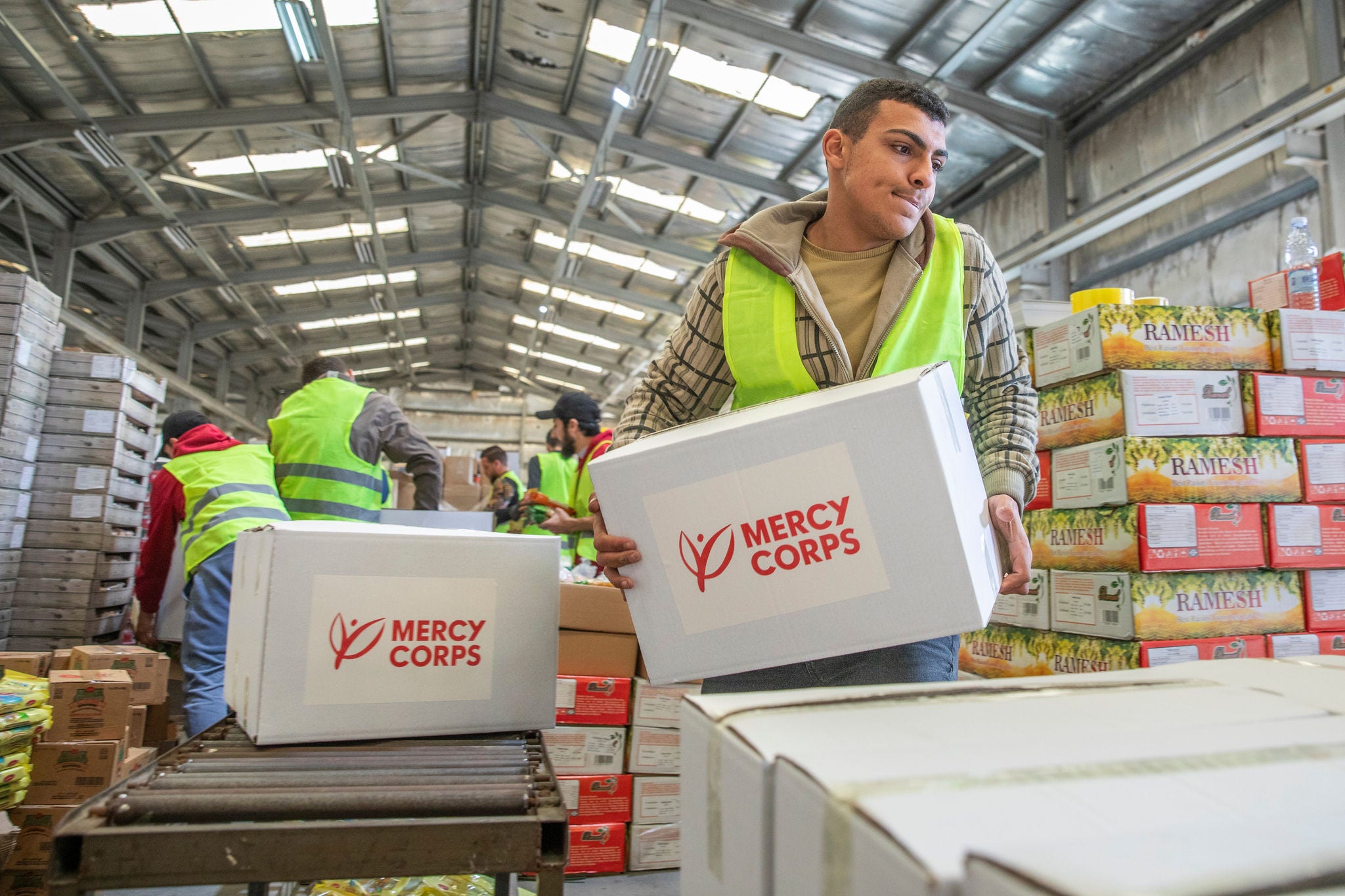 Gemini said A warehouse worker in a neon safety vest carries a large white box labeled with the Mercy Corps logo, with other workers and stacks of supplies in the background.