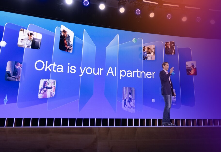 Todd McKinnon, CEO and Co-Founder of Okta, speaks on stage at the Oktane conference in front of a large screen that reads, "Okta is your AI partner."