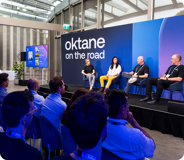 Four panelists seated on a stage in front of a blue backdrop with the text "Oktane on the road" speaking to an audience.