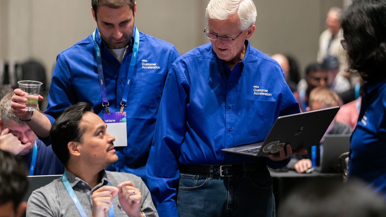 Two Okta Customer Acceleration team members in blue shirts help an attendee on a laptop at the Oktane conference.