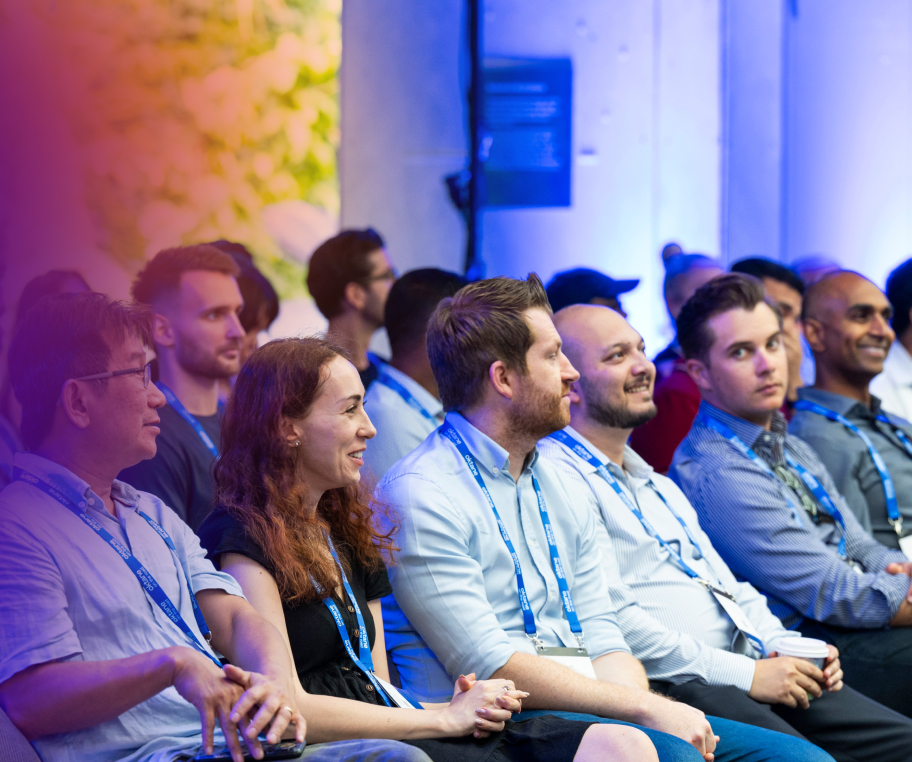A group of conference attendees wearing blue lanyards sitting in rows and attentively listening to a presentation.