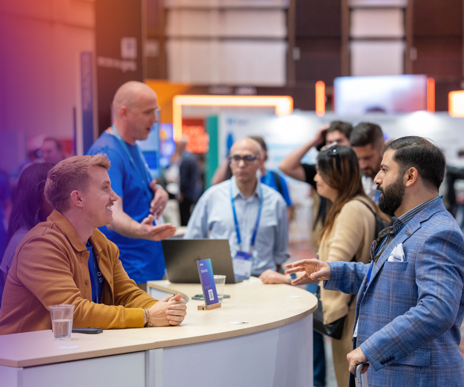 A man in a blue plaid blazer conversing with staff seated behind a curved white reception counter during a busy tech conference.