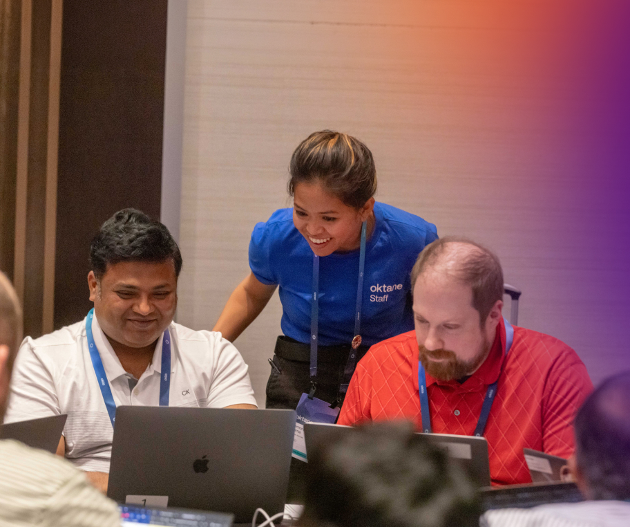 A woman wearing a blue "Oktane Staff" shirt smiling and helping two men sitting at laptops during a training session.
