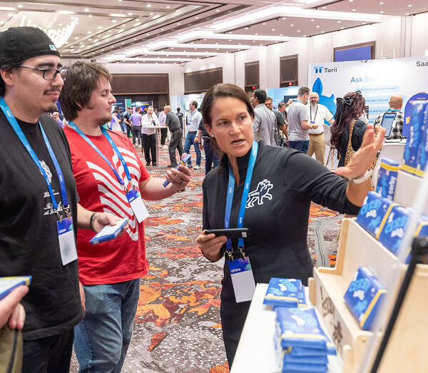  A female exhibitor shows branded merchandise to two male attendees at a booth in the Oktane expo hall.
