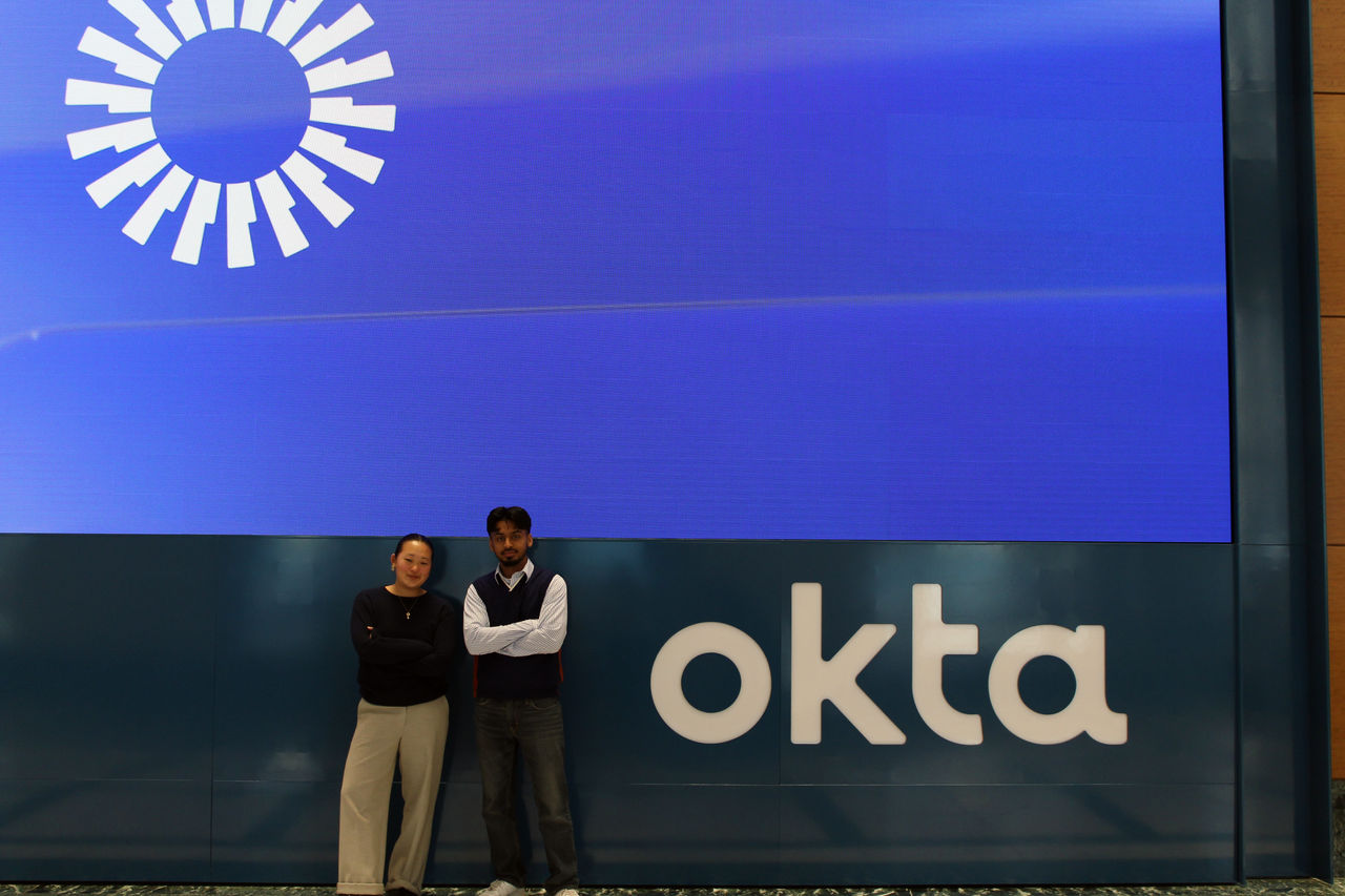 Two individuals, a man and a woman, stand in front of a large blue wall featuring the Okta logo and a circular graphic design. 