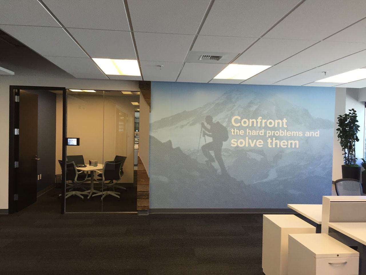 Modern office interior at Okta's Bellevue office with a mountain wall graphic reading 'Confront the hard problems and solve them' next to a glass conference room