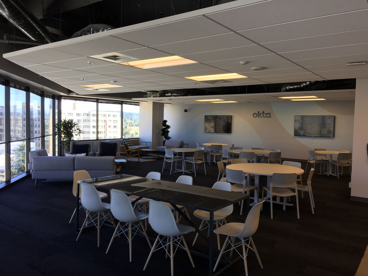 Spacious open lounge and seating area in Okta's Bellevue office with long tables, round dining sets, window seating, and the Okta logo on the wall