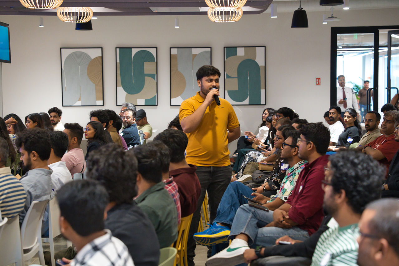 A man in a yellow shirt stands in the middle of a seated audience, holding a microphone to ask a question.