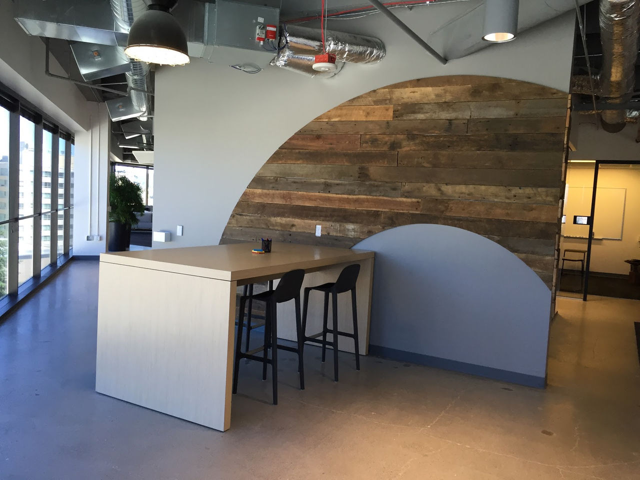 Modern office lounge in Okta's Bellevue office with a high-top wooden table, bar stools, large floor-to-ceiling windows, and a reclaimed wood accent wall