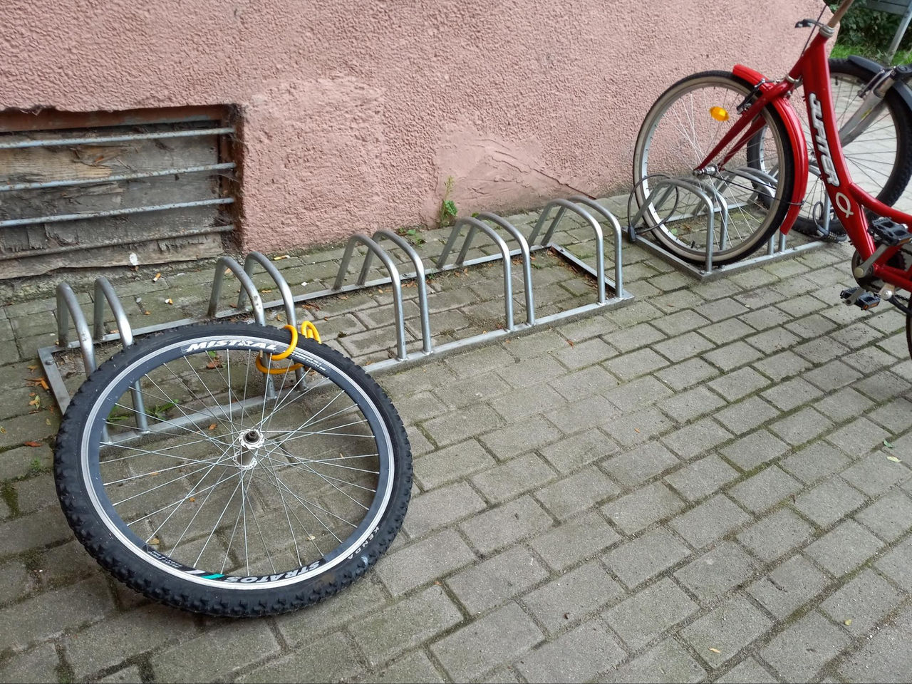 A detached bicycle wheel lies on the ground next to a metal bike rack on a paved sidewalk. 