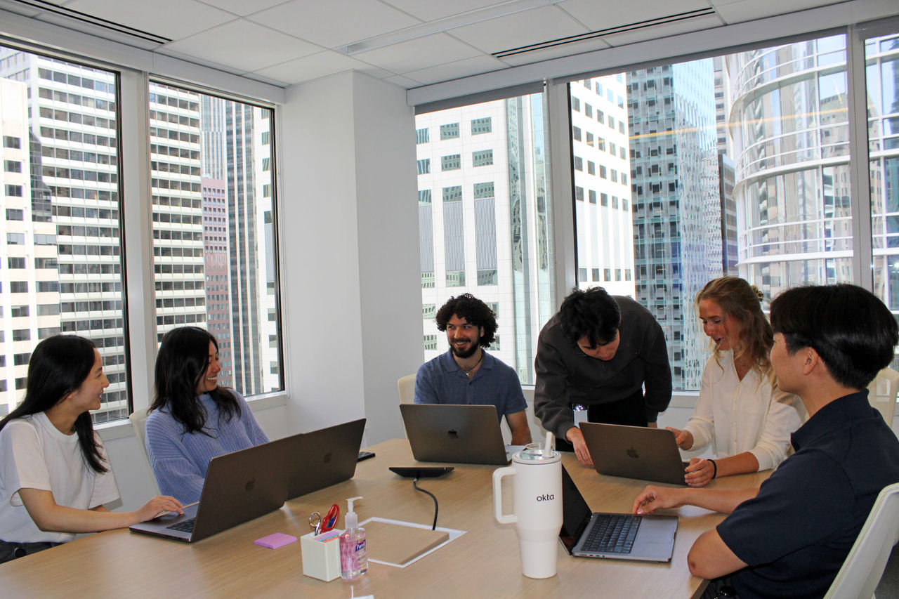 A group of professionals is gathered around a table in a bright, modern office space with large windows showcasing urban skyscrapers.