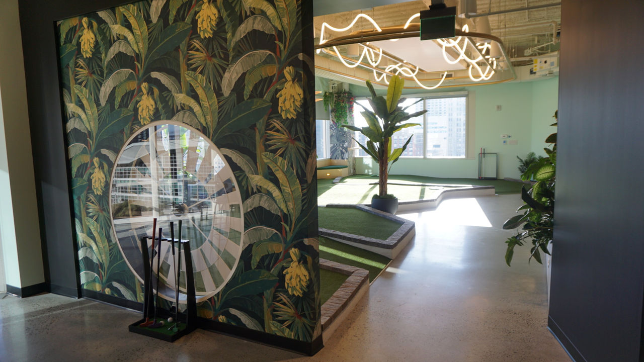 A vibrant indoor mini-golf area featuring tropical-themed wall decor with leafy patterns and a circular mirror. 