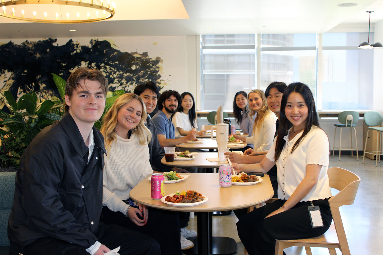 A group of individuals seated at round tables in a bright, modern office cafeteria. 