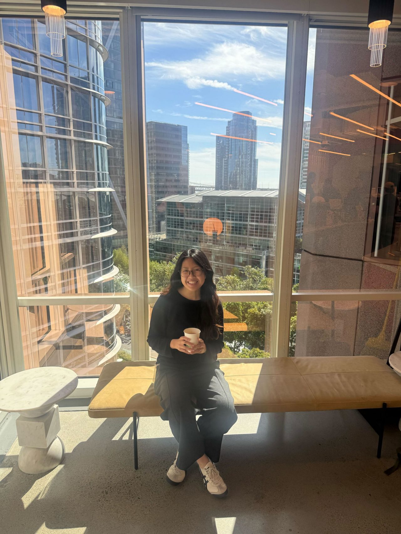 A person sits on a wooden bench holding a coffee cup in a modern indoor space.