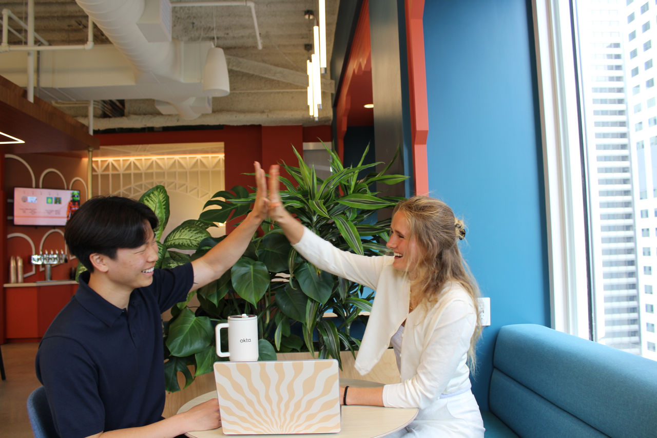 Two individuals are seated at a round table in a contemporary office space, engaging in a high-five gesture. 